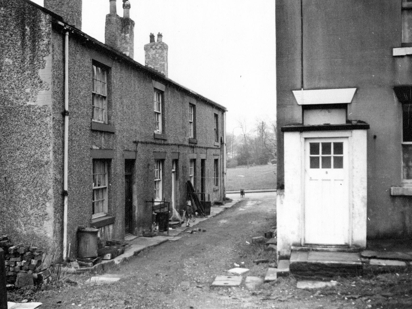 A view looking on to Mill Lane. Unmade access to houses on the left. Some are back-to-back while the one actually on Mill Lane is a through terrace. A small boy and a dog can be seen outside the houses.