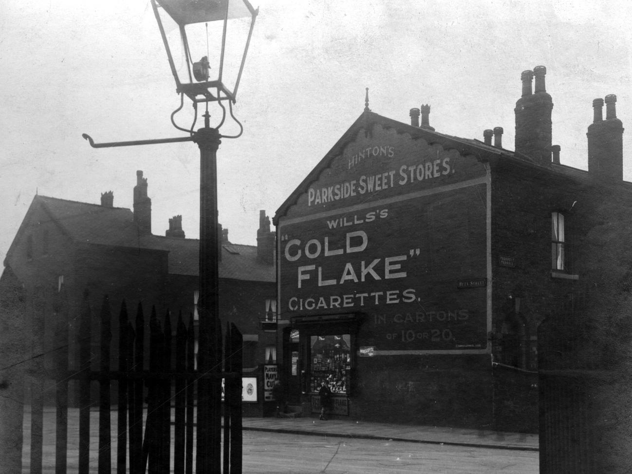 This photo shows Hunslet Moor railway crossing on Beza Street. Number 48 Parkside sweet stores is opposite with a large advertisement for Wills Gold Flake cigarettes above. Prospect Street and Prospect Terrace are partially visible