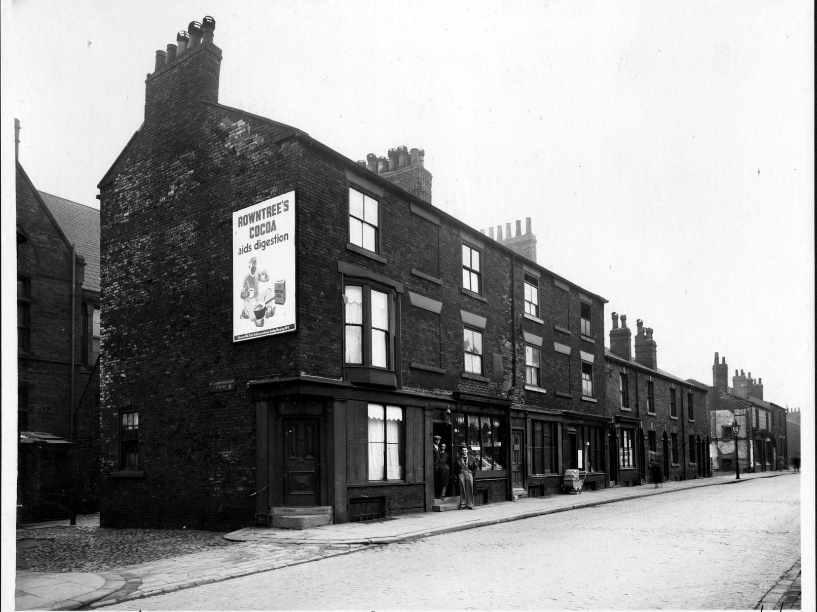 Accommodation Road in Burmantofts. There is a gap where the Spinners Arms public house had been demolished. A pram is outside the doctors. A poster for Rowntree's Cocoa on the end wall.