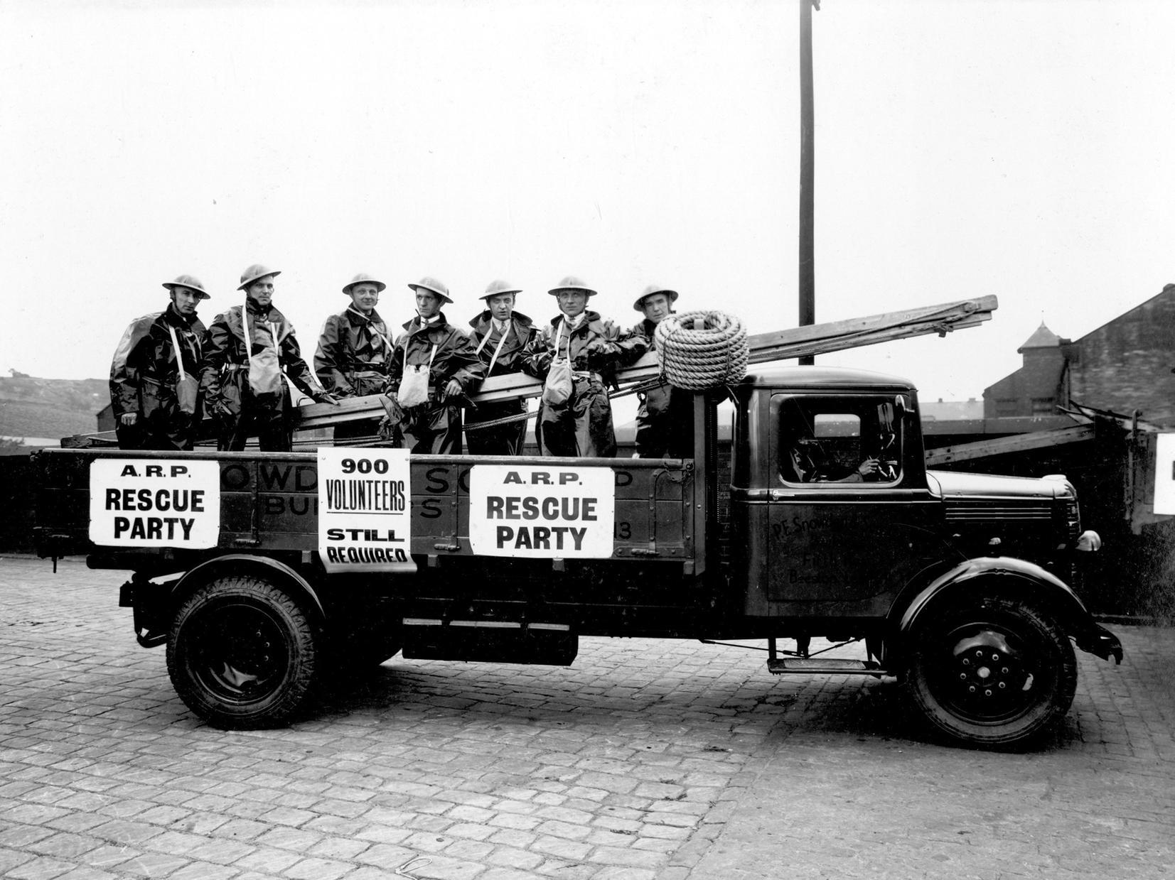 A.R.P. (Air Raid Precaution) rescue party stand on the back of a Bedford truck, equipped with ladders, rope and other rescue equipment. The men are dressed in protective clothing with tin hats and gas mask bags.