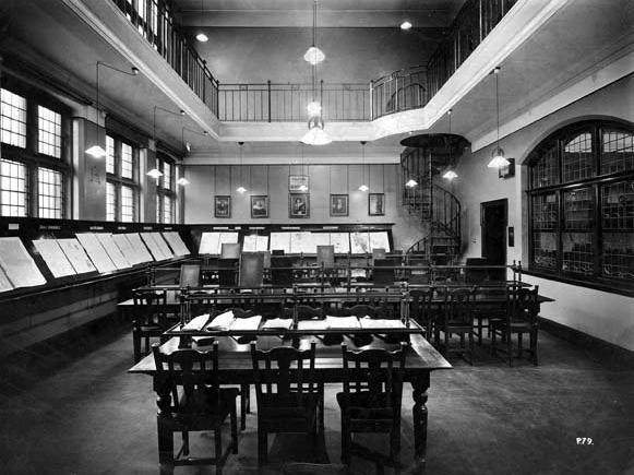 The newsroom at Chapel Allerton Library. The papers are fixed to reading rests around the room. The tables have rests for magazines. A spiral staircase in the right corner of the room gives access to the balcony.