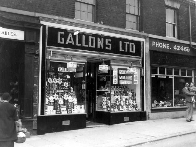 Gallons Ltd was a grocers on Harrogate Road with a double fronted window showcasing its wares. On the right is TW Smith Florist and a man holding a young child is stood on the pavement.