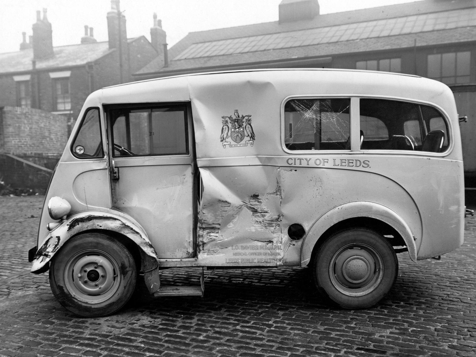 A Leeds Public Health Department ambulance, a Morris van, after an accident, parked in a Leeds City Transport garage on York Road. There is a clear view of the Leeds coat of arms on the bodywork.