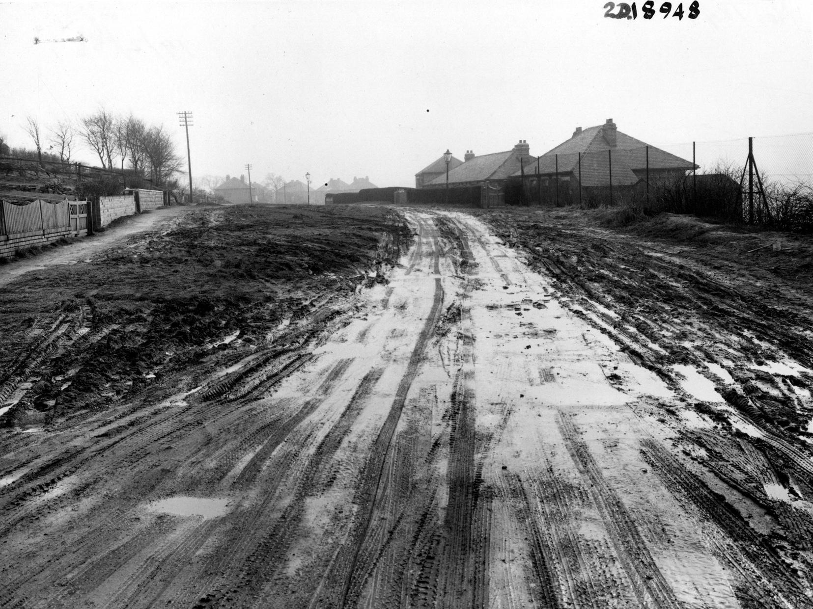Blue Hill Lane in Wortley from near the junction with Tong Road. The picture shows the unmade, muddy surface with tyre tracks. The roofs of houses are visible on the right, beyond a wire mesh fence.