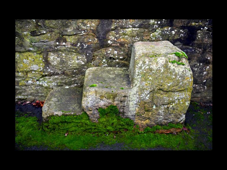 Mounting block, 18th century or earlier. Sandstone block cut to make 3 steps, set against wall of churchyard.
